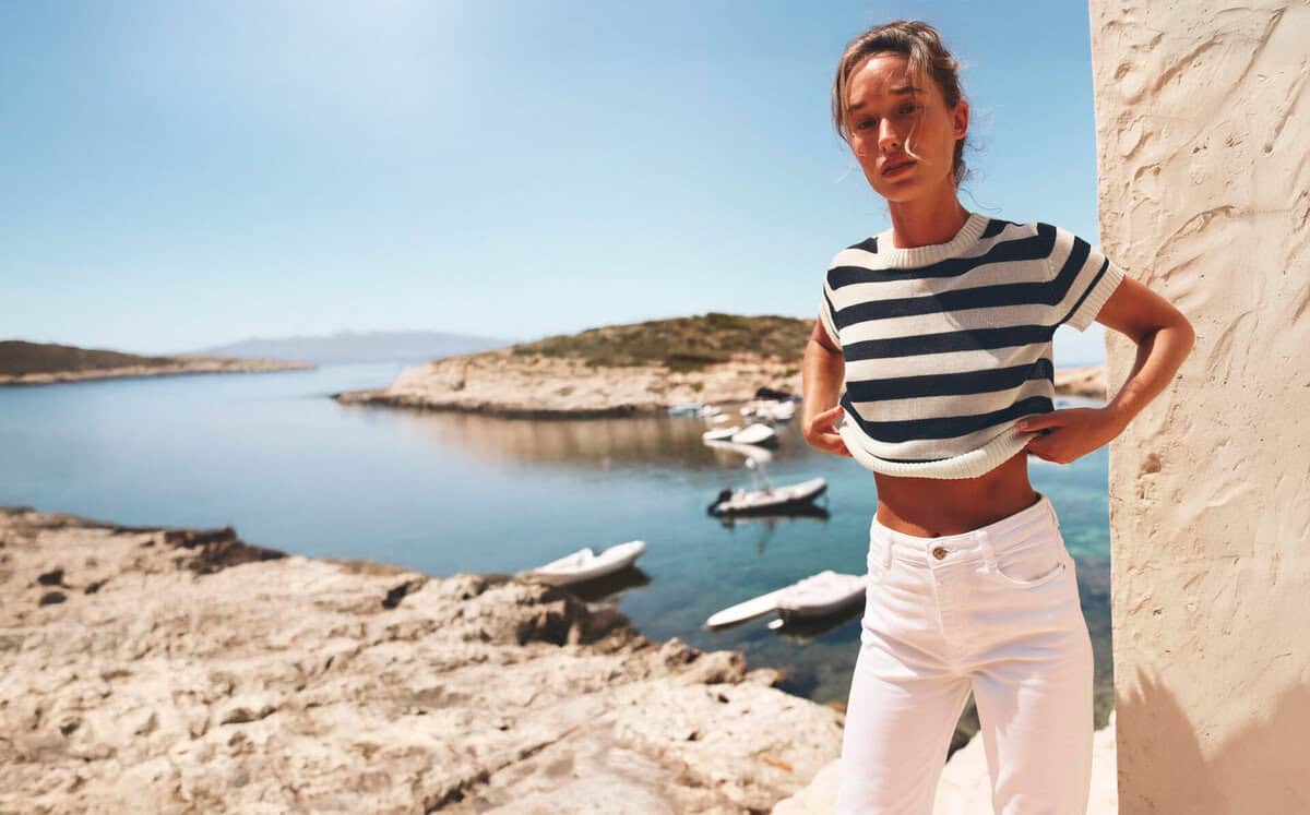 Una donna con una maglia a righe e pantaloni bianchi in piedi vicino all'acqua.